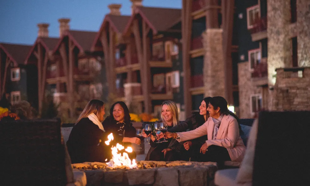 Girlfriends sit around firepit on Fire & Water Terrace and cheers with wine.