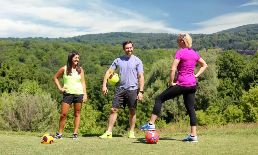 Three people standing on a golf course with soccer balls ready to play foot golf