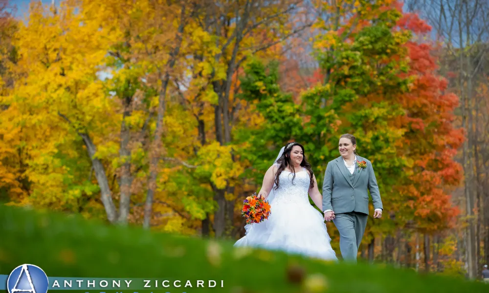 Bride and bride walking hand in hand with fall trees behind them.