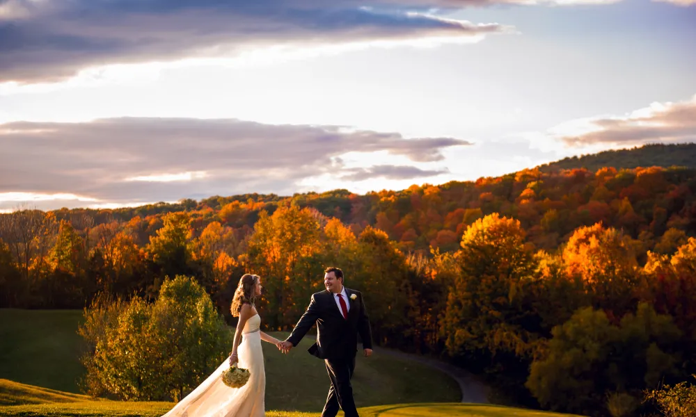 Bride and groom walking hand in hand with fall foliage behind them.