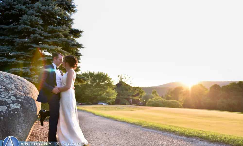 Bride and groom lean against a large rock as sun sets behind them.