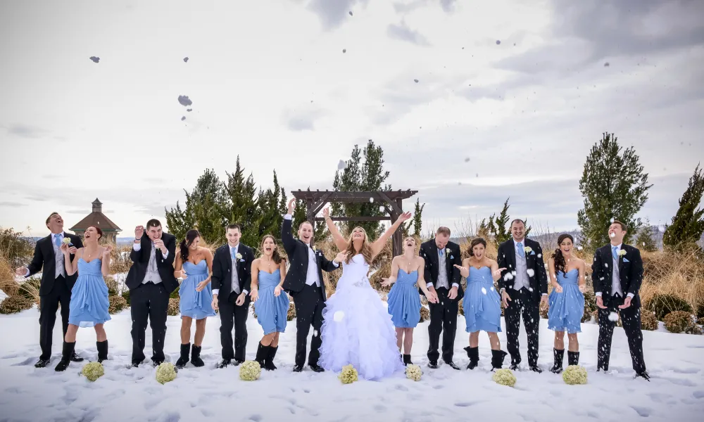 Wedding party, featuring men in black tuxes and bridesmaids in short blue dresses, stand together in a line and throw snow in air.