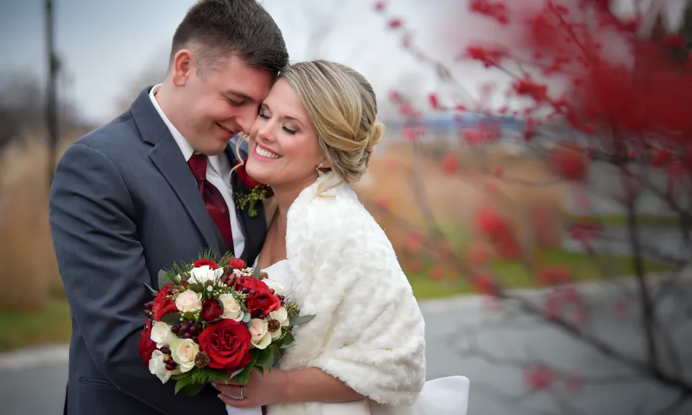 Bride and groom stand together with eye closed as bride holds bouquet of white and red roses.