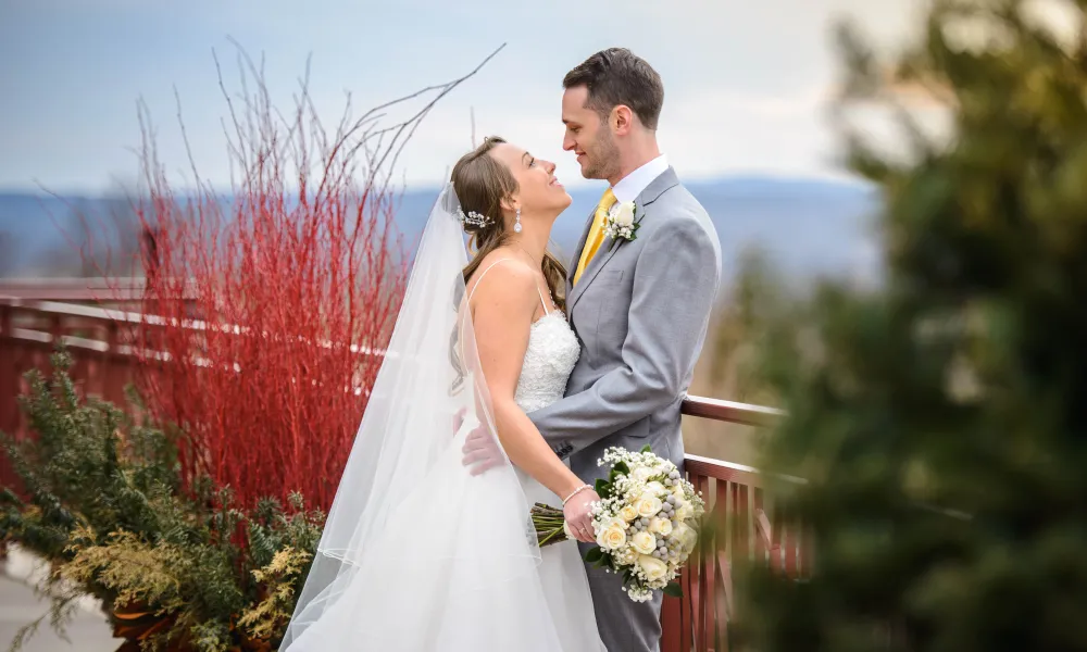 Bride and groom look at one another as they lean on railing with mountains behind them.