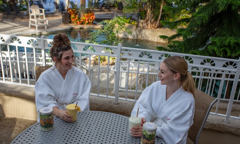 Two women sitting in white robes at a table in the Biosphere eating lunch and drinking cocktails.