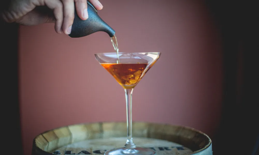 Hand pouring cocktail into martini glass. on top of a barrel.