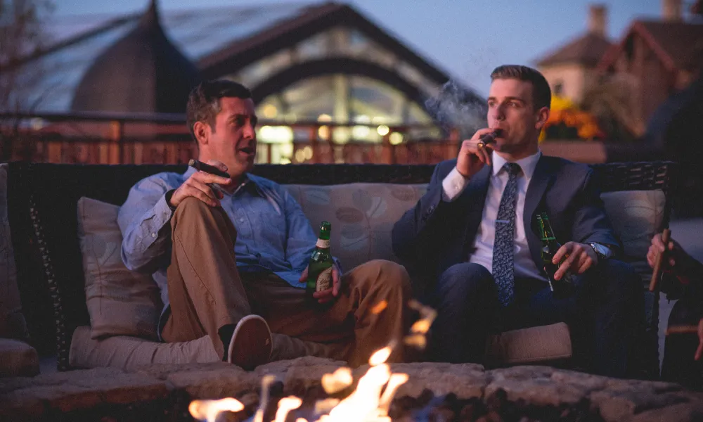 Two guys smoking cigars by a fire pit