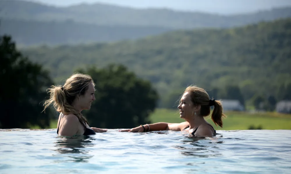 Two women speaking to one another as they relax in the vista 180 pool with mountains in background.