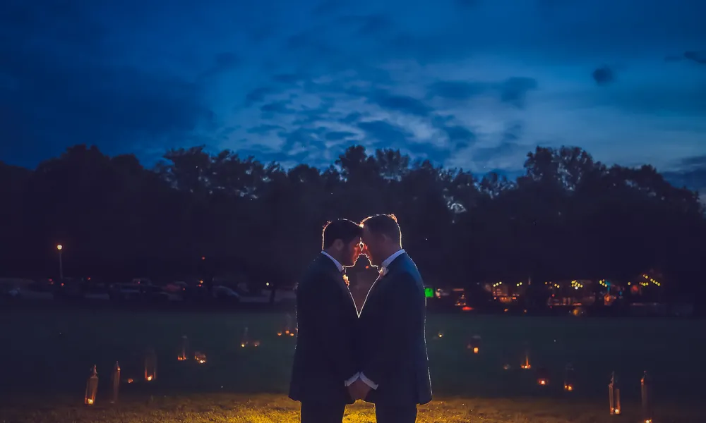 Grooms with foreheads placed together standing in a candle lit walkway.