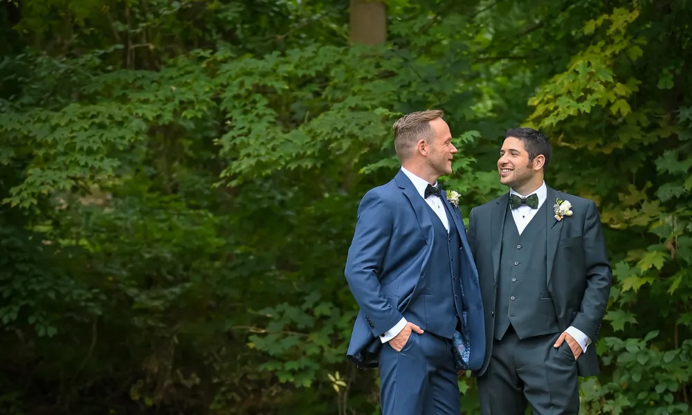 Grooms, wearing blue suits, looking at one another while standing in front of green foliage.