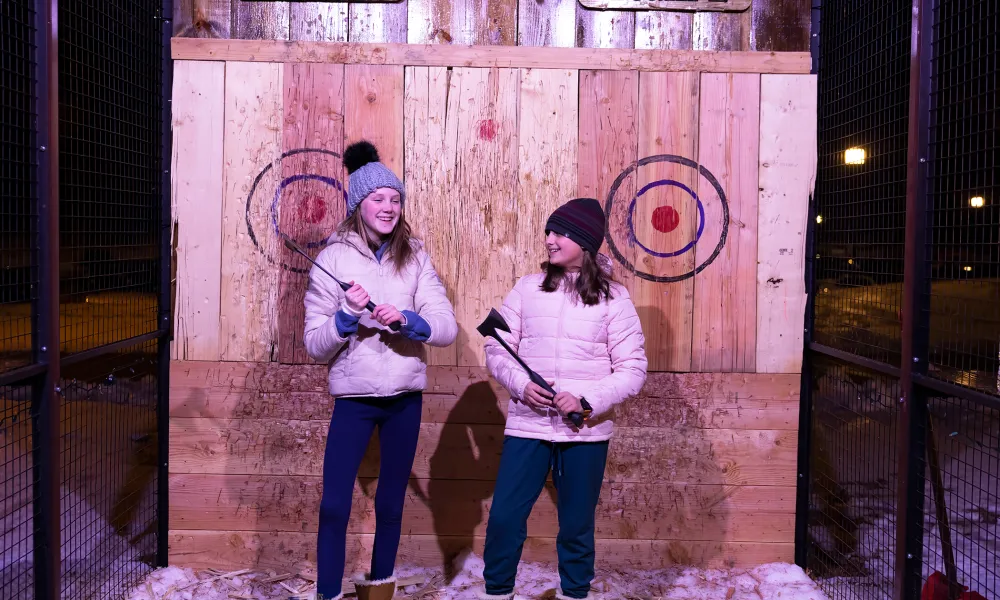 Two girls wearing white winter jackets and holding axes in their hands to throw.