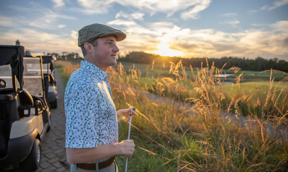 Man stands near golf carts holding club with sun setting in background.