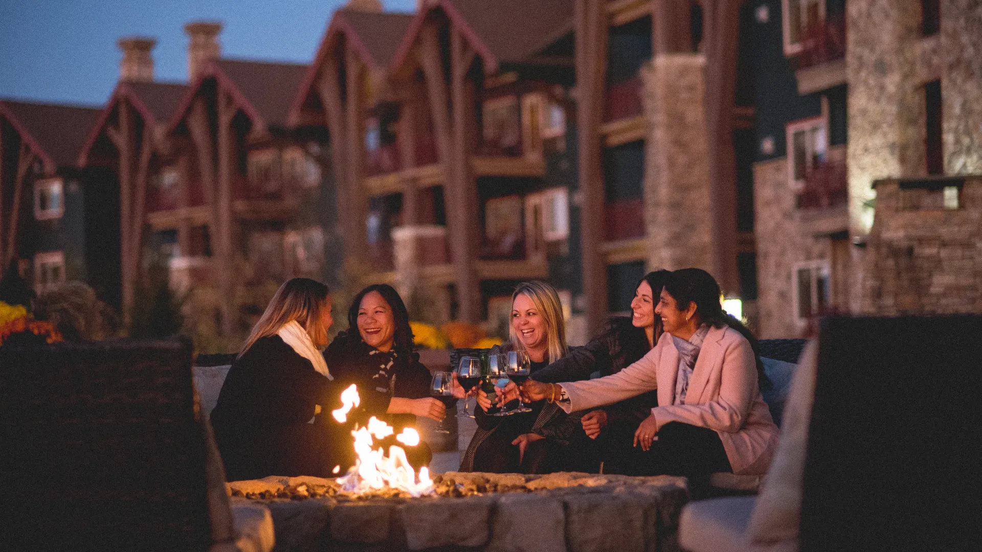 Girlfriends sit around firepit on Fire & Water Terrace and cheers with wine.