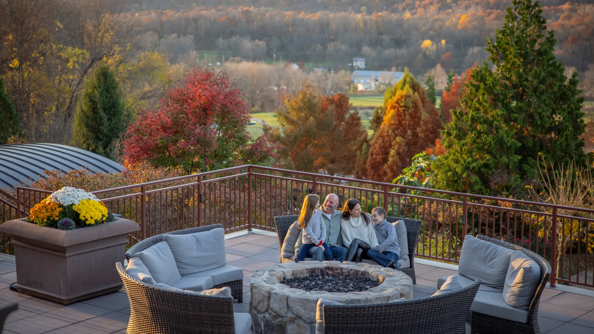 Family of 4 sitting by firepit on fire &amp; water terrace with fall background.