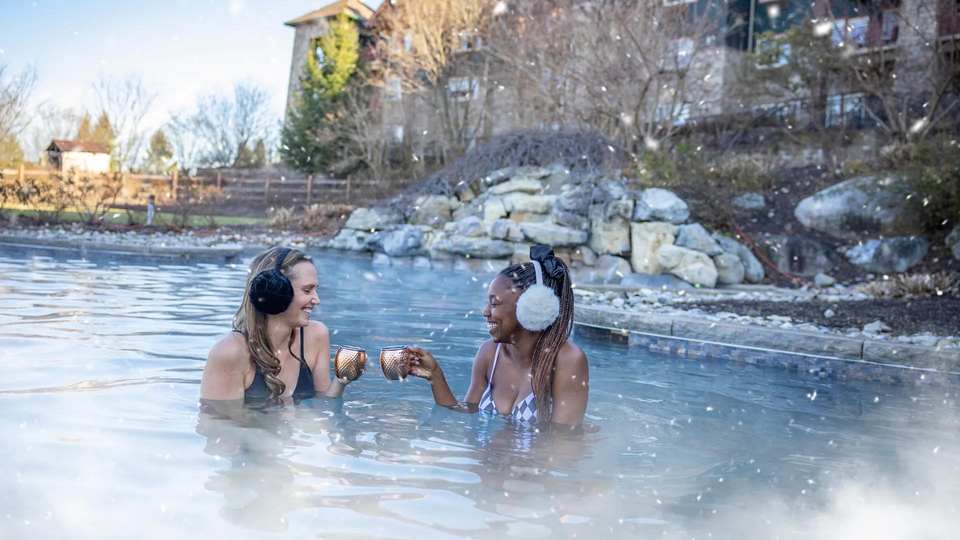 Two women in outdoor snowpool wearing earmuffs and drinking mules.