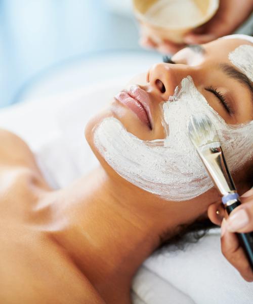 Tech painting white face mask on woman's face during spa treatment.