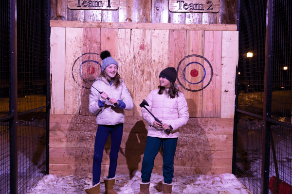 Two girls wearing white winter jackets and holding axes in their hands to throw.