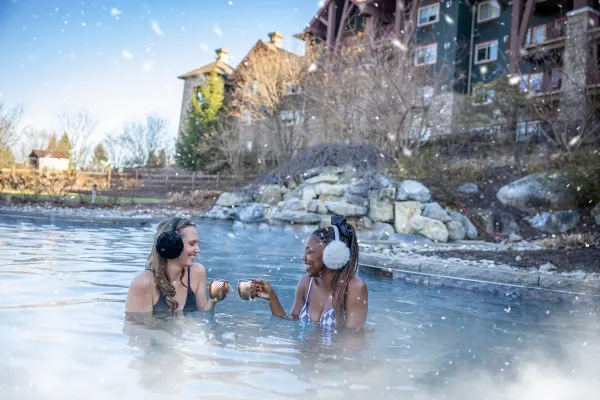 Two women in outdoor snowpool wearing earmuffs and drinking mules.