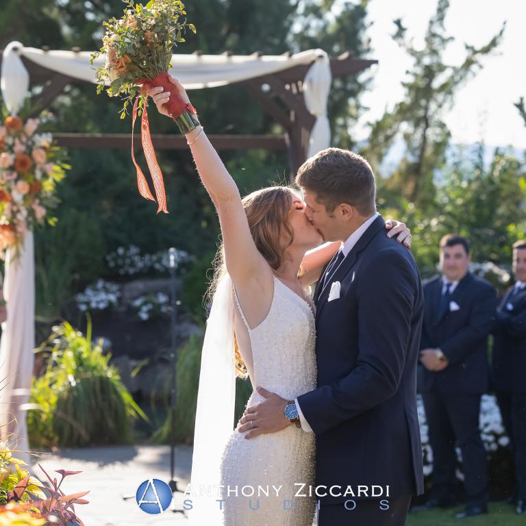 Bride and groom kiss under arch during their Bride walking down the aisle at Wedding Garden ceremony space at Crystal Springs Country Club. 