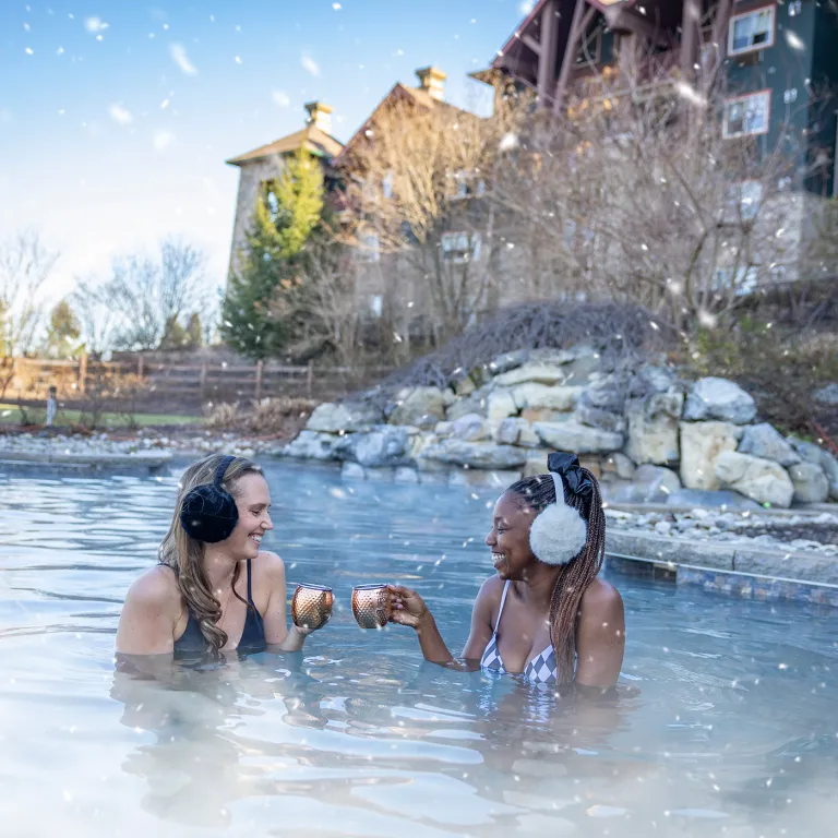 Two women in outdoor snowpool wearing earmuffs and drinking mules.